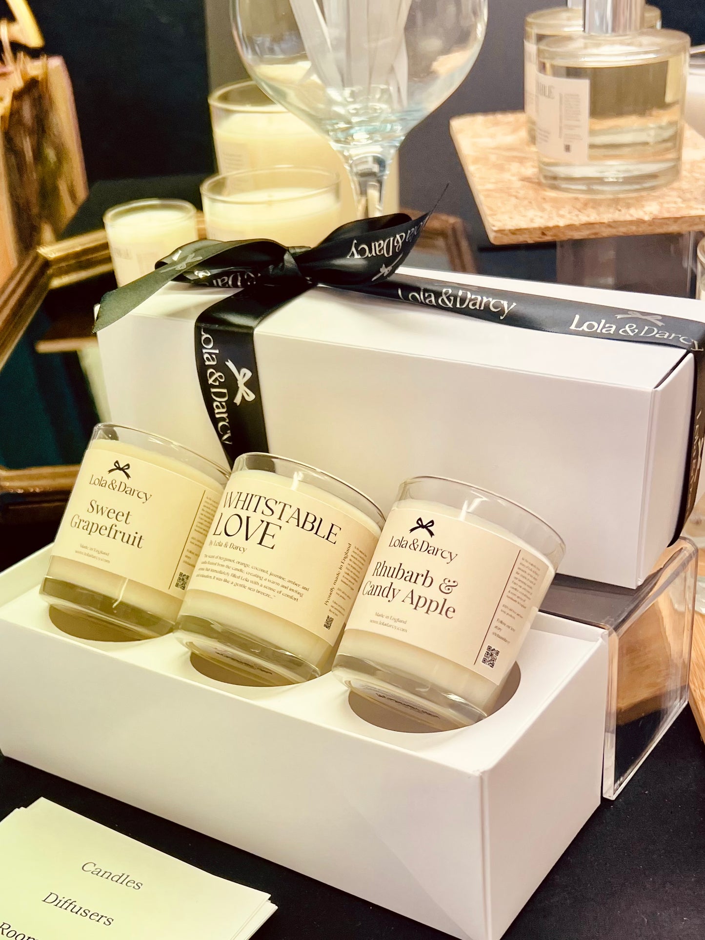 A trio of candles in beige glass jars, each labeled with a different scent, presented in a white box with a black ribbon. The background includes a candle holder and a mirror.