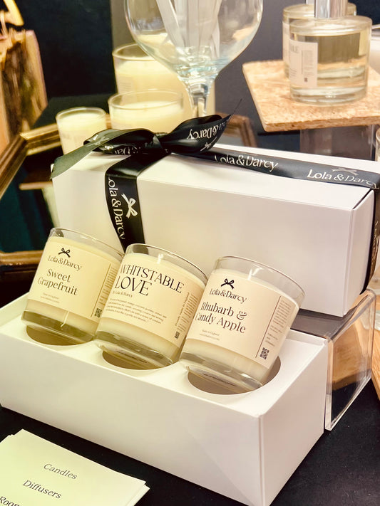 A trio of candles in beige glass jars, each labeled with a different scent, presented in a white box with a black ribbon. The background includes a candle holder and a mirror.