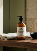 Bottle of 'Whitstable Beach' soap on a wooden surface with a towel and bowl in a softly lit room.